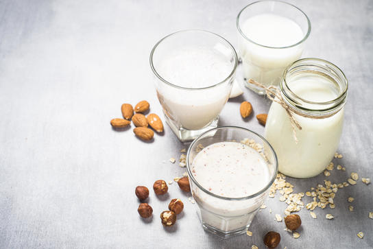 Four glasses of dairy and non dairy beverages surrounded by almonds, hazelnut and oat.