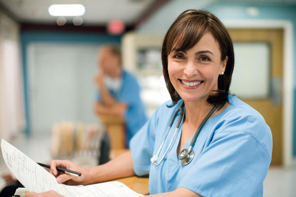 Femme médecin portant un uniforme bleu et un stéthoscope autour du cou. Elle tient un stylo et du papier. Elle sourit.